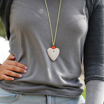 Close-up of a handmade necklace featuring a white heart-shaped lava stone pendant, accented with an orange glass bead, on a light green suede cord.